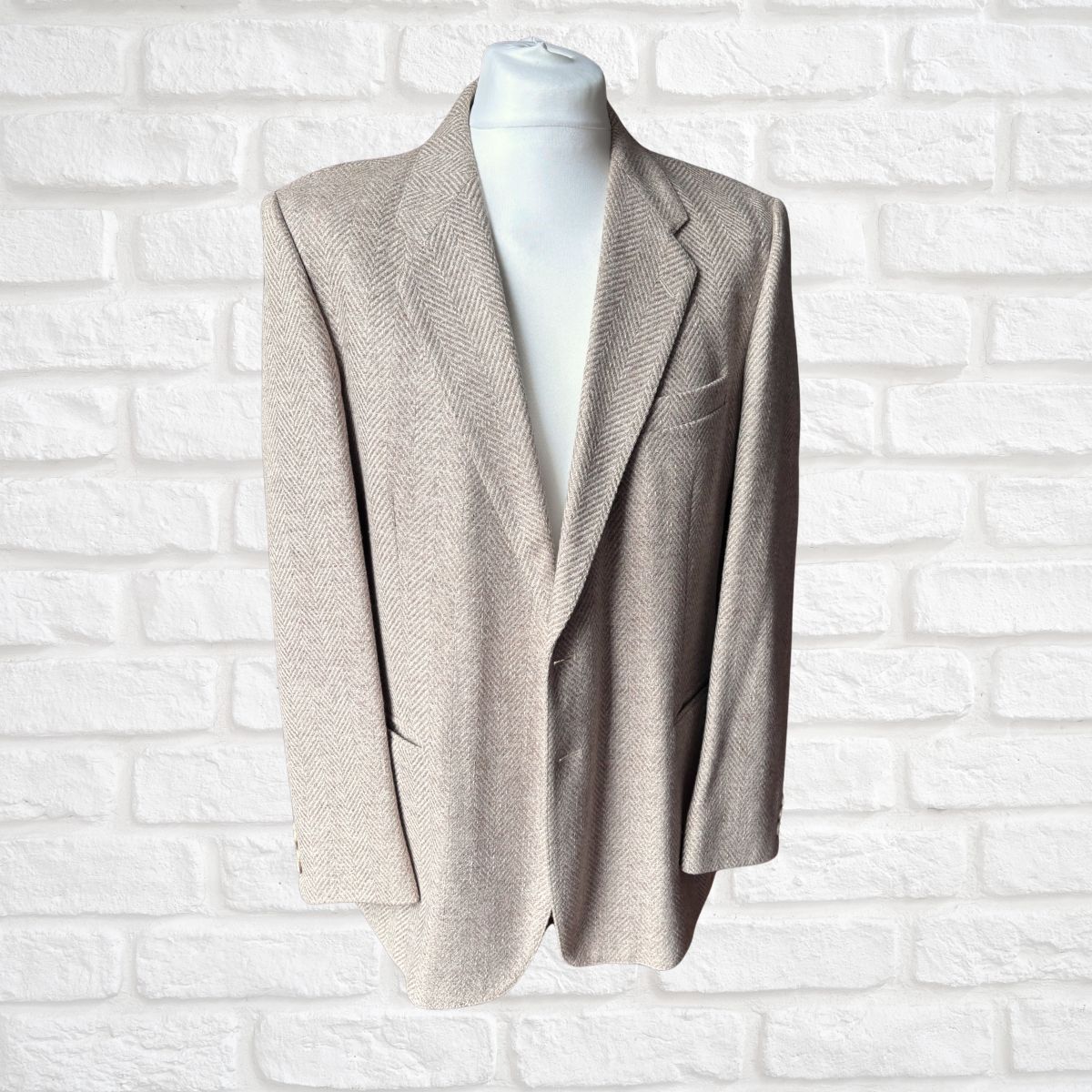 A men's single-breasted blazer in a light beige herringbone pattern, displayed on a white mannequin torso against a white painted brick wall background.