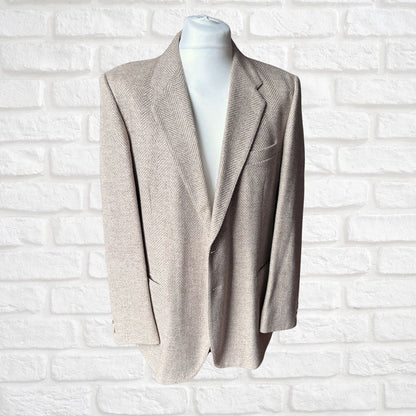 A men's single-breasted blazer in a light beige herringbone pattern, displayed on a white mannequin torso against a white painted brick wall background.