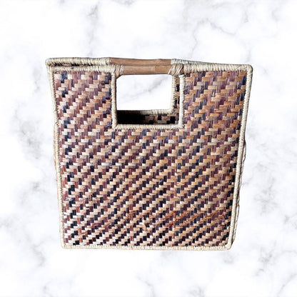 A square woven basket bag with a cutout handle, featuring a textured chevron pattern in brown, beige, and muted purple tones, set against a white marble background.