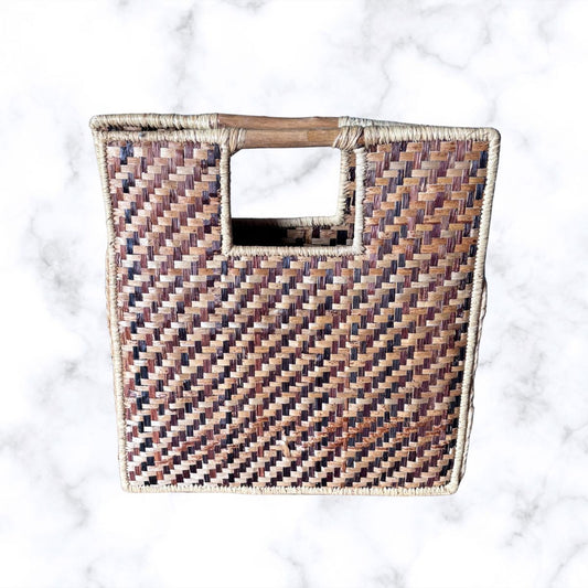 A square woven basket bag with a cutout handle, featuring a textured chevron pattern in brown, beige, and muted purple tones, set against a white marble background.