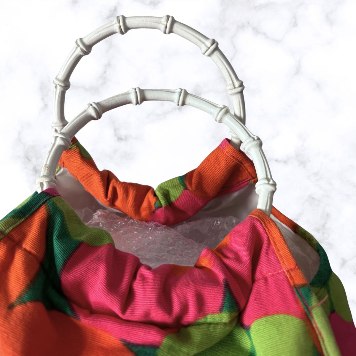 A close-up of a colorful fabric bag with a bright pink, orange, and green pattern, featuring a gathered fabric neck trim and a white faux-bamboo style handle, set against a white marble background.