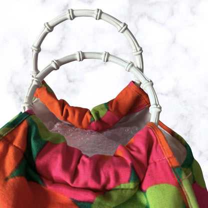 A close-up of a colorful fabric bag with a bright pink, orange, and green pattern, featuring a gathered fabric neck trim and a white faux-bamboo style handle, set against a white marble background.