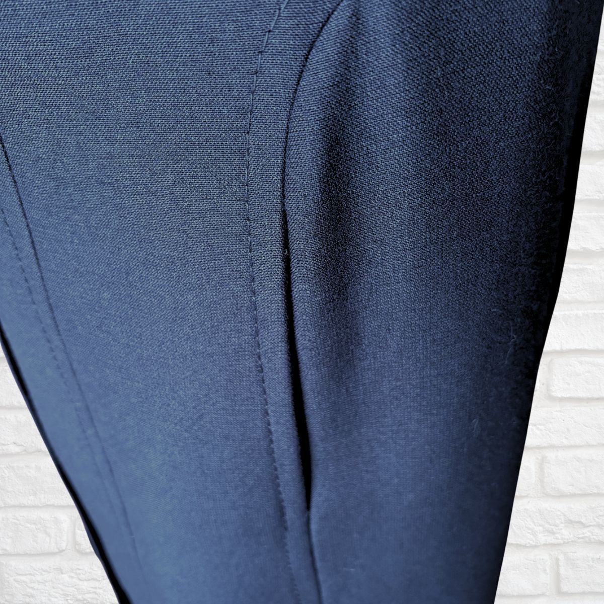 Close-up of navy blue fabric pocket with visible stitching and a textured weave, shown against a white brick wall background.