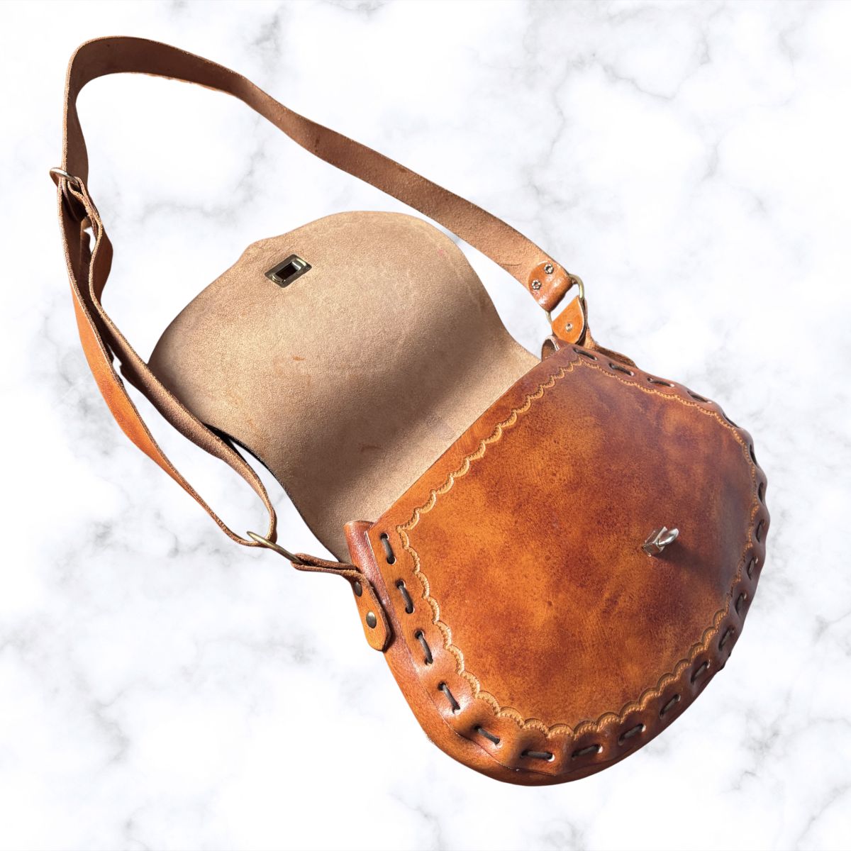 A brown leather shoulder bag with a curved, saddle-like shape, featuring a tan interior lining, decorative scalloped stitching, and a matching adjustable shoulder strap, displayed on a white marble background.
