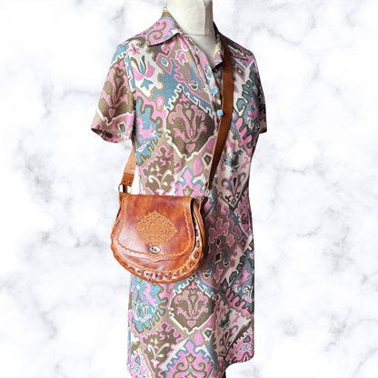 A brown leather crossbody bag with a decorative floral tooling design on the front flap, displayed on a mannequin wearing a short-sleeve patterned shirt dress. The bag has a brown strap and a silver-tone snap button closure.