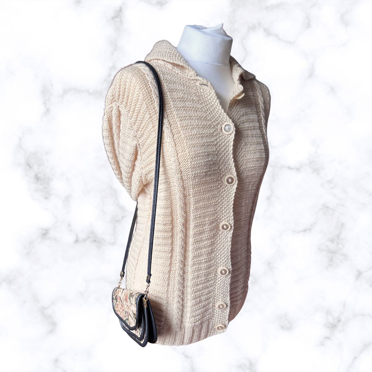 A cream-colored, textured knit cardigan sweater displayed on a white mannequin torso, styled with a small black shoulder bag against a white marble background.