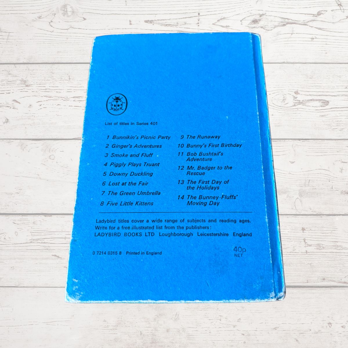 A blue children's book cover or back cover photographed on a light whitewashed wooden surface. The cover has printed track/listing text and a small circular logo near the top left.