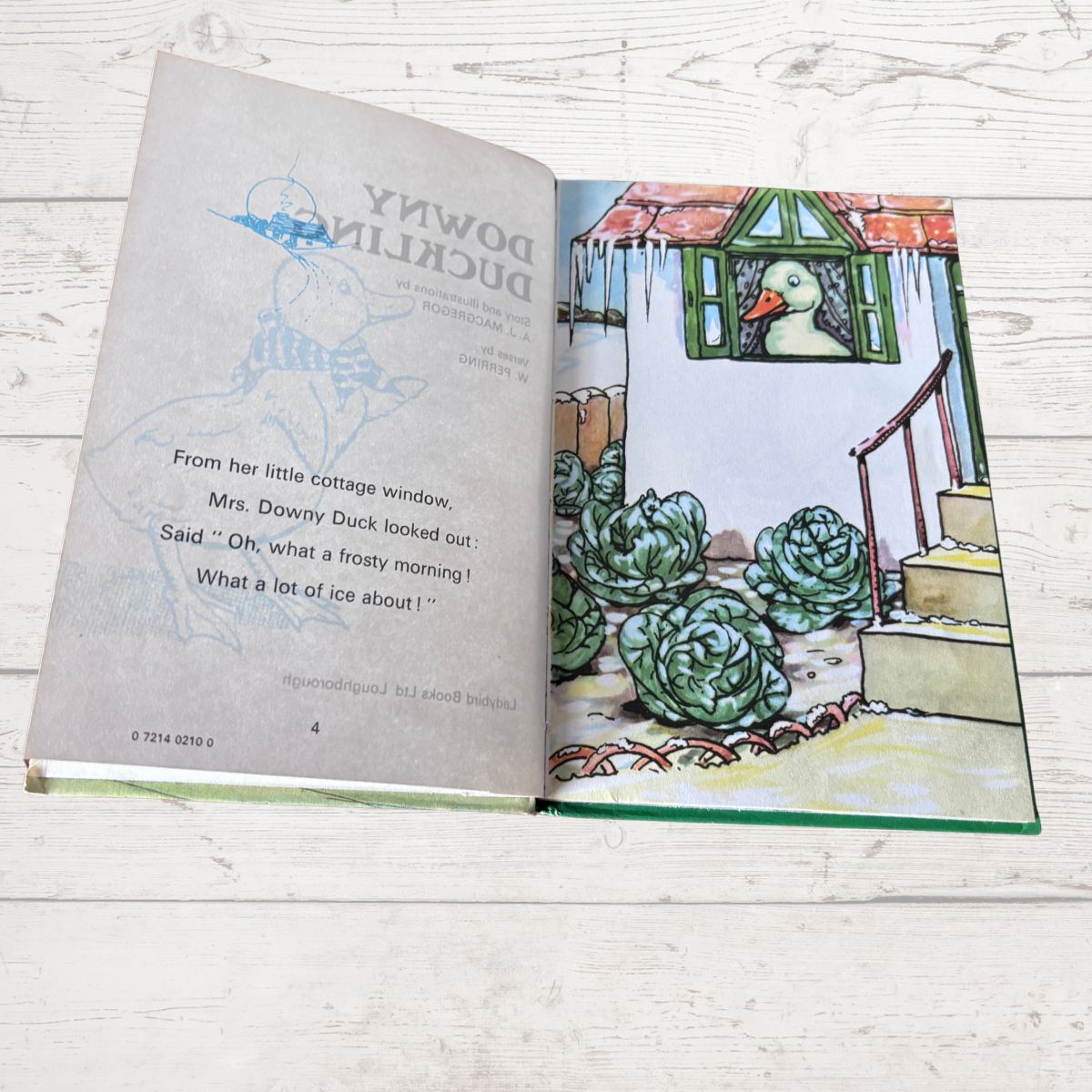 An open children's book titled 'Downy Duckling' laid flat on a white wood surface. The left page shows the title and a light blue duck illustration, while the right page features a colorful illustration of a duck in a cottage garden with cabbage-like plants.