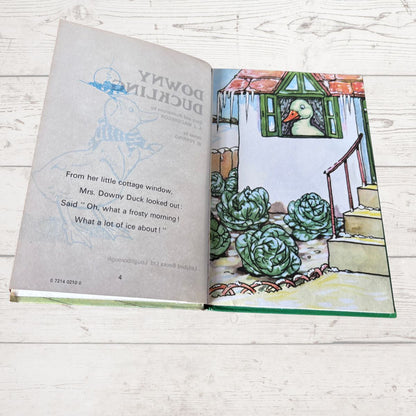 An open children's book titled 'Downy Duckling' laid flat on a white wood surface. The left page shows the title and a light blue duck illustration, while the right page features a colorful illustration of a duck in a cottage garden with cabbage-like plants.