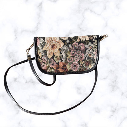 A small shoulder bag with a black leather trim and a floral tapestry pattern in pink, beige, and green, featuring a long black leather strap on a white marble background.