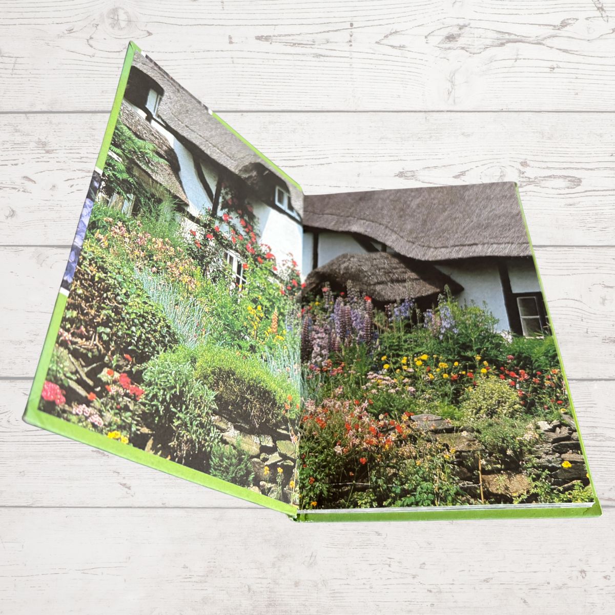 An open hardcover book lying on a light whitewashed wooden surface. The pages show a printed photograph of a thatched cottage surrounded by colorful flower gardens.