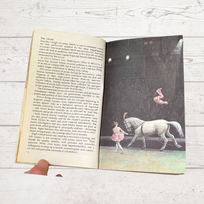 An open vintage children's book lying on a white wood surface, showing printed text on the left page and a black-and-white illustration of a girl riding a horse on the right page.