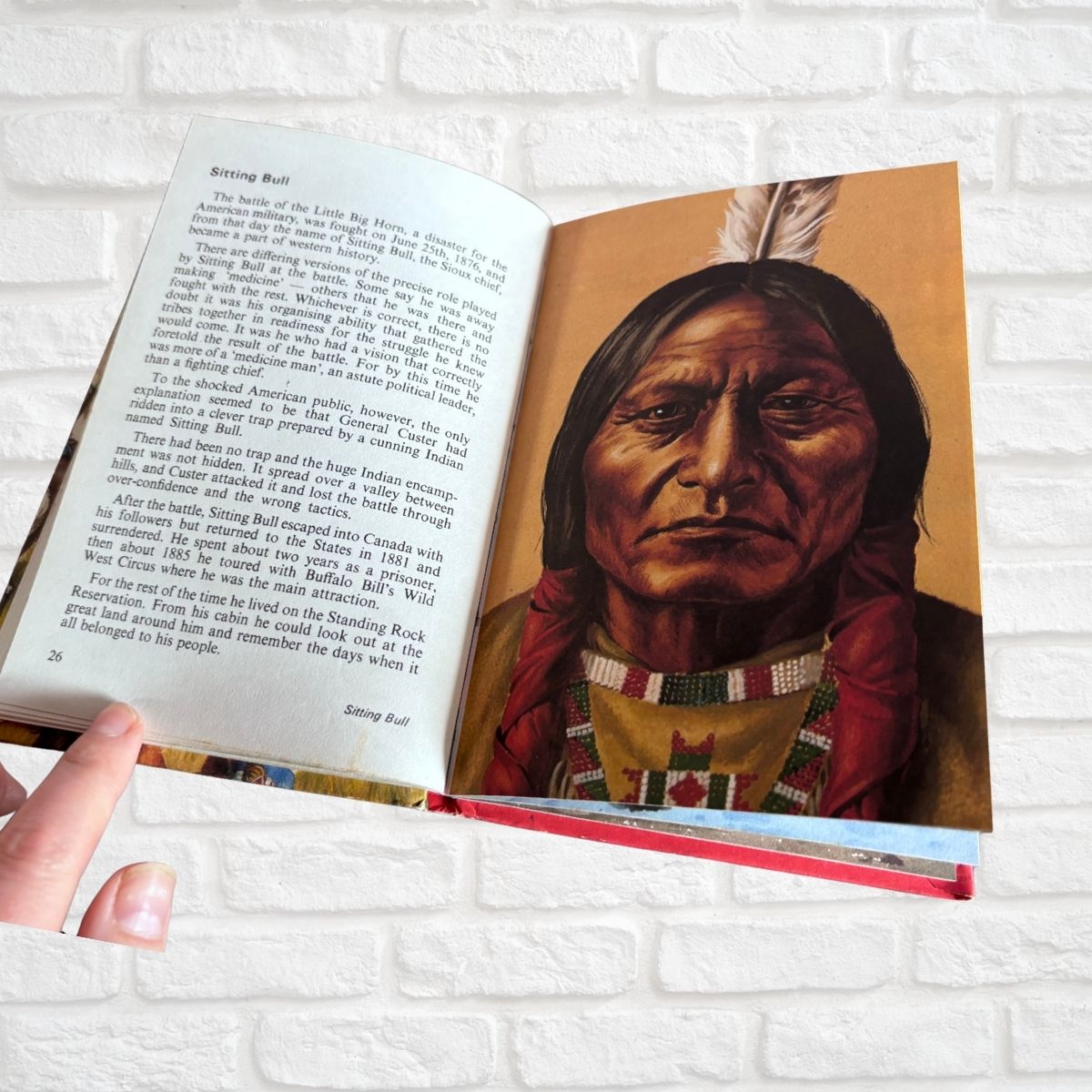 An open book showing a printed page with text on the left and a painted portrait of Sitting Bull on the right, held by a hand against a white brick wall background.