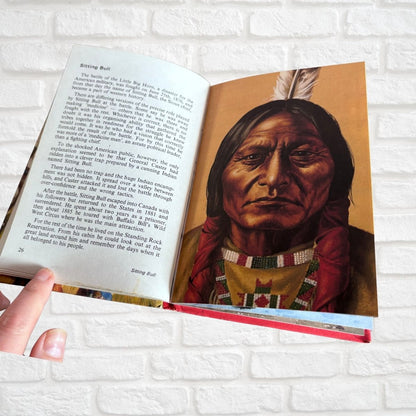 An open book showing a printed page with text on the left and a painted portrait of Sitting Bull on the right, held by a hand against a white brick wall background.