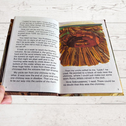 An open book showing printed text on the left page and a colorful map of a volcanic landscape on the right page, held by a hand over a white wood background.