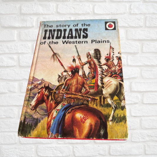 Vintage children's book titled 'The story of the Indians of the Western Plains' leaning against a white painted brick wall. The cover features colorful illustrated Native American riders on horseback.