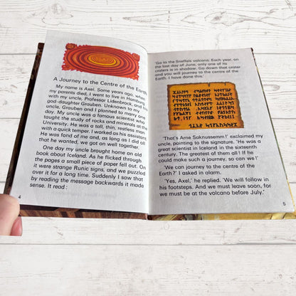 A hand holds an open book showing two printed pages with text and a small orange map illustration. The background is a white painted wood surface.