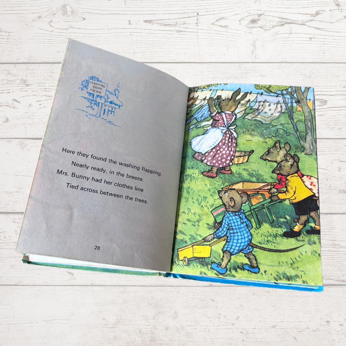 An open children's book lying on a light wood surface, showing a gray left page with text and a colorful right page with an illustrated scene of animals in a garden.