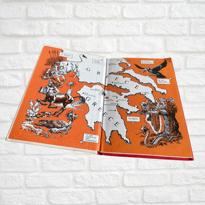 An open vintage-style illustrated map book showing Greece, photographed against a white brick wall background.