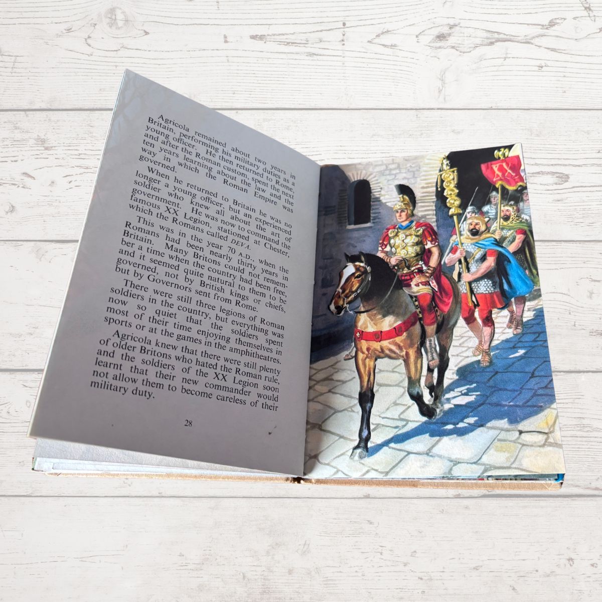 An open book lying on a white wooden surface, showing printed text on one page and a colorful illustration on the other of soldiers on horseback in historical dress.
