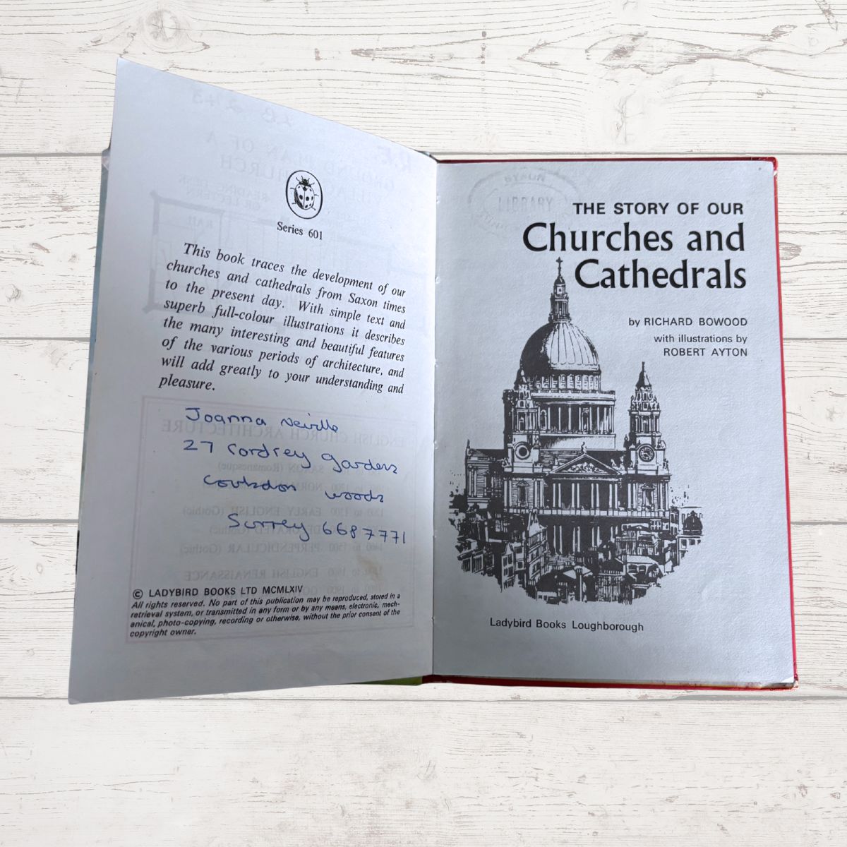 An open book titled 'The Story of Our Churches and Cathedrals' by Richard Bowood, showing the front cover illustration of a cathedral and the handwritten address 'Joanna Munsie 27 Contrey Gardens, Luton, Bedfordshire' on the inner page, photographed on a light wood surface.
