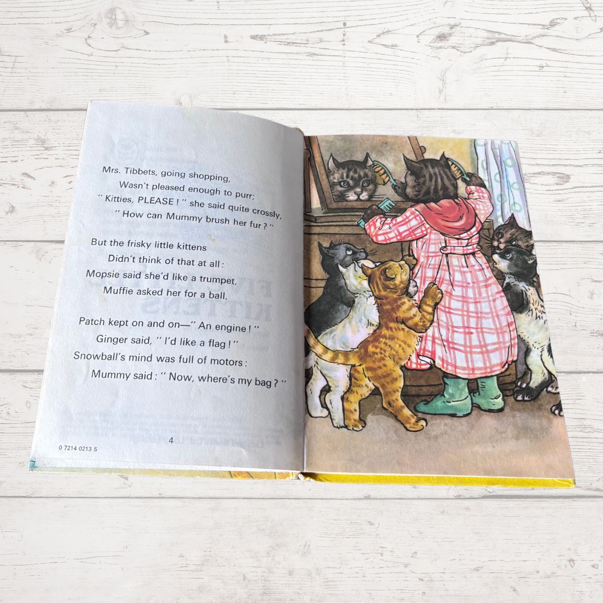 An open children's book lying on a white wood surface, showing printed text on the left page and a colorful illustration on the right page of a girl in a pink plaid dress interacting with several cats.