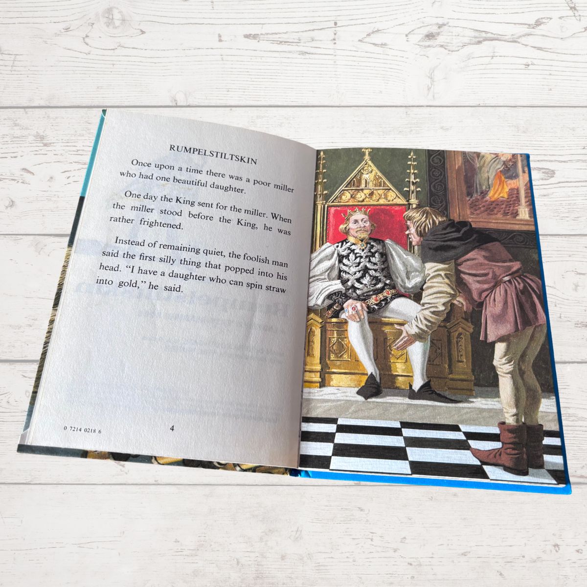 An open children's book lying on a white wood surface, showing a printed story page with text on the left and an illustrated scene on the right of a king in a throne room with a woman and a man.