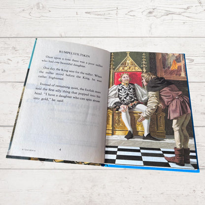 An open children's book lying on a white wood surface, showing a printed story page with text on the left and an illustrated scene on the right of a king in a throne room with a woman and a man.