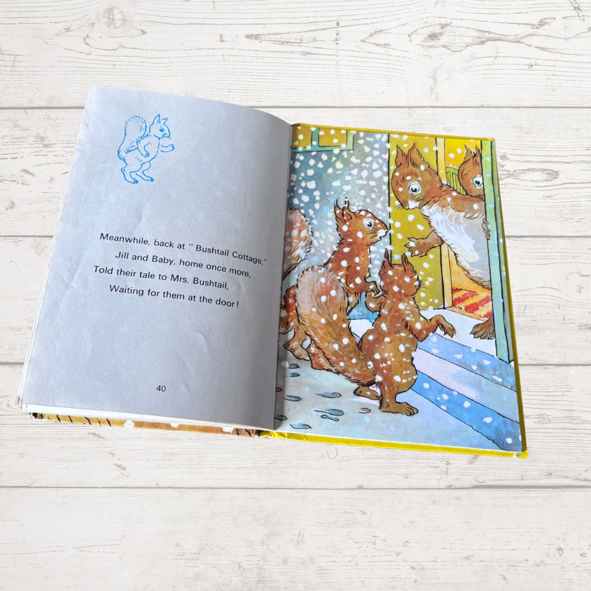 An open children's book lying on a light wood surface, showing a printed page with text on the left and a colorful illustration of squirrels on the right.