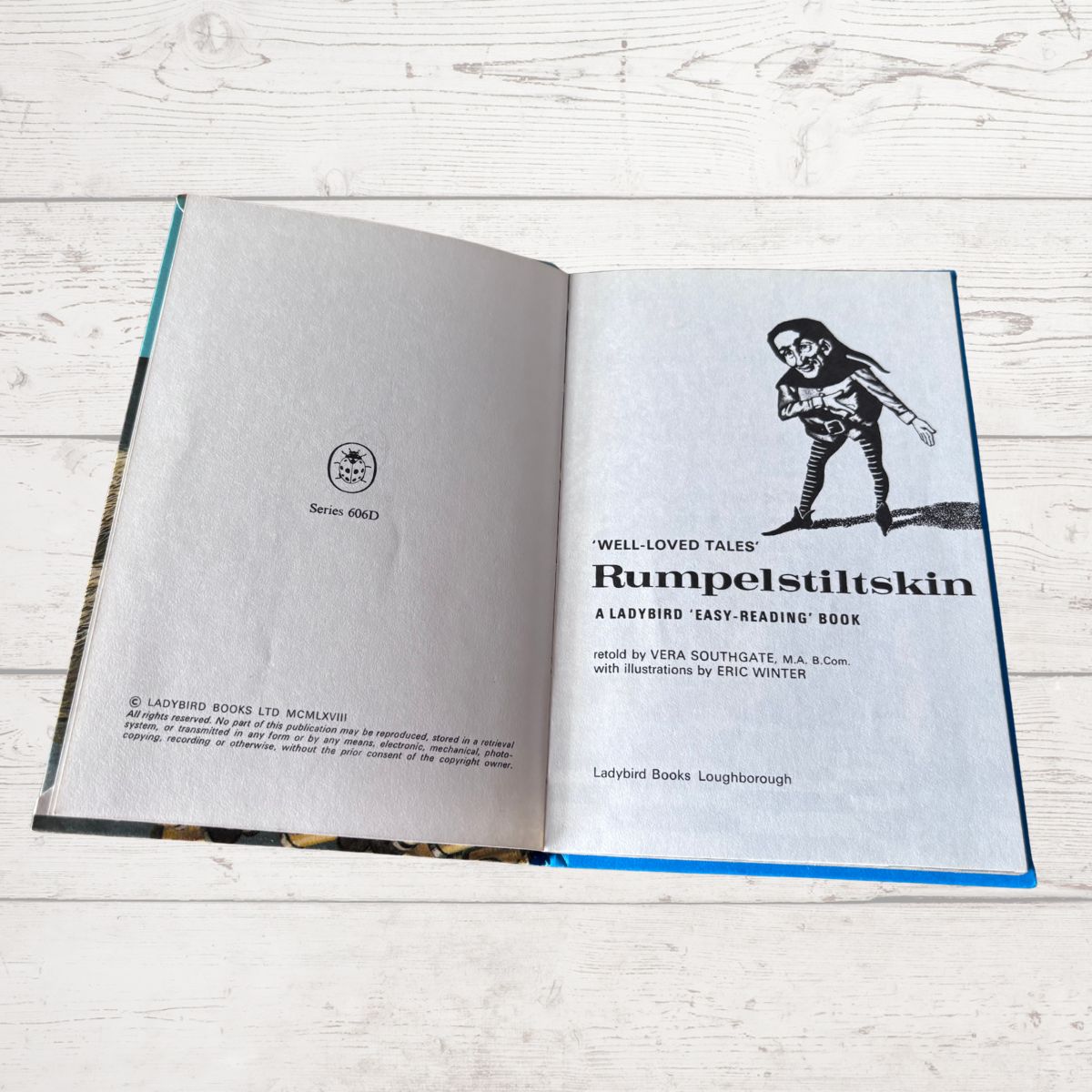 An open book titled 'Rumpelstiltskin' from 'Well-Loved Tales' by Ladybird Books, displayed on a light-colored wooden plank background. The book's pages are off-white with black text and a black-and-white illustration of a skeleton figure.
