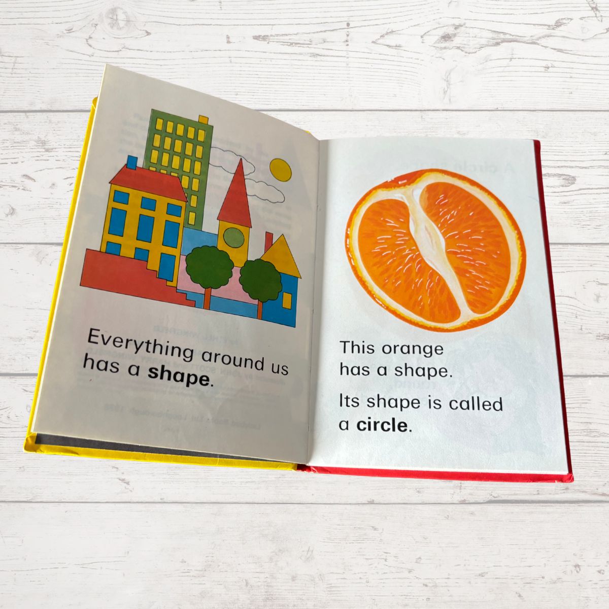Open book showing illustrations of a cityscape and an orange with text about shapes on a white wooden surface.