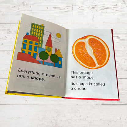 Open book showing illustrations of a cityscape and an orange with text about shapes on a white wooden surface.