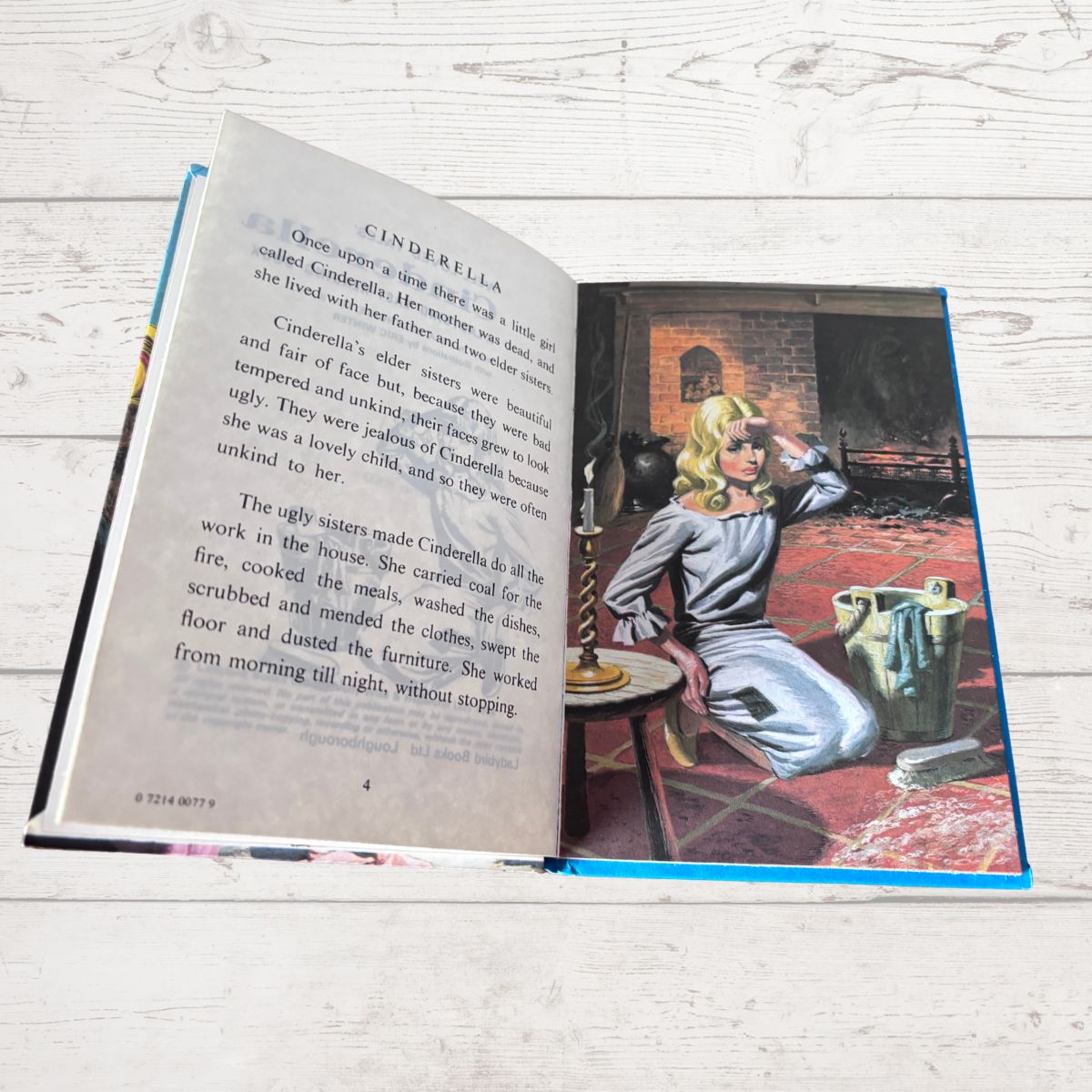 An open hardcover book titled Cinderella, photographed on a light whitewashed wooden surface. The left page contains printed text, and the right page shows an illustration of Cinderella in a gray gown sitting in a dark room with a mirror and broom.