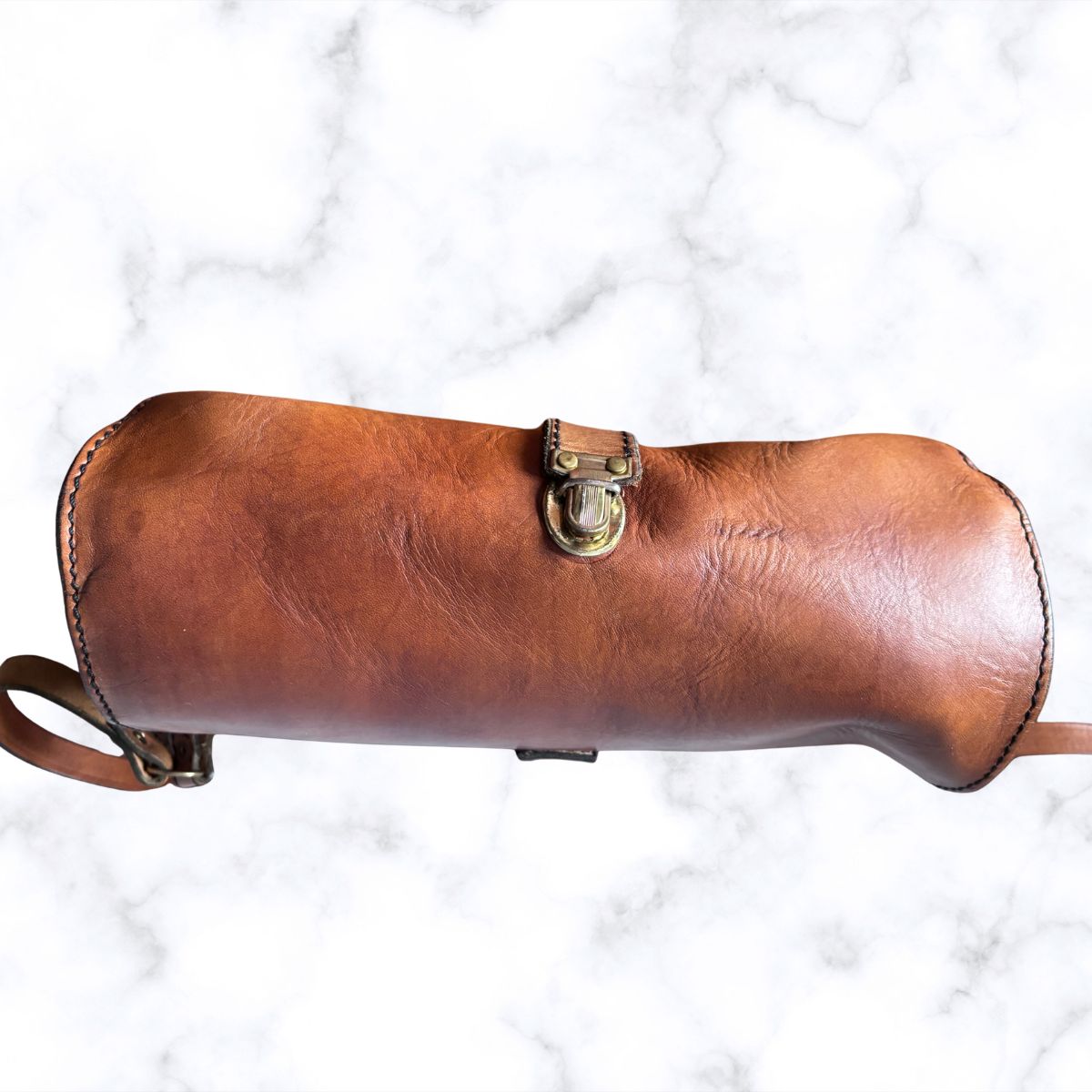 A brown leather handbag with a brass-tone clasp closure, photographed horizontally against a white marble-patterned background.