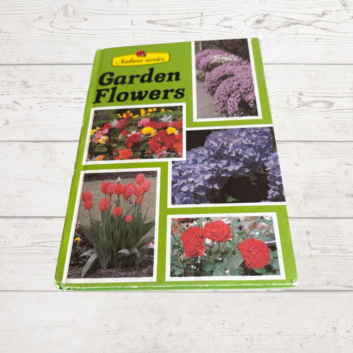 A hardcover gardening book titled 'Garden Flowers' from the 'nature series' lies on a white-washed wooden surface. The book cover features a bright green border with five square color photographs showcasing different garden flowers, including red roses, red tulips, purple hydrangeas, and yellow/orange flowers.