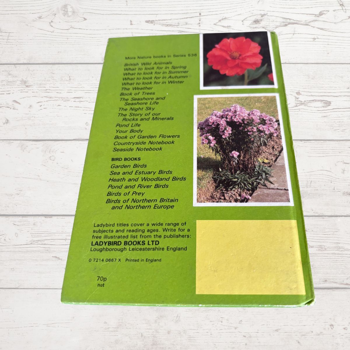 The back cover of a green Ladybird Books showing the title list, publisher details, and two small inset photos of a red flower and purple flowers, photographed on a white wood background.