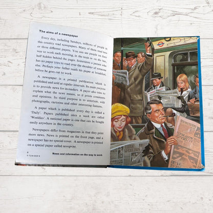 An open children's book titled 'The aims of a newspaper' displayed on a light gray wood background. The left page contains black text, and the right page features a colorful cartoon illustration of people holding and reading newspapers.