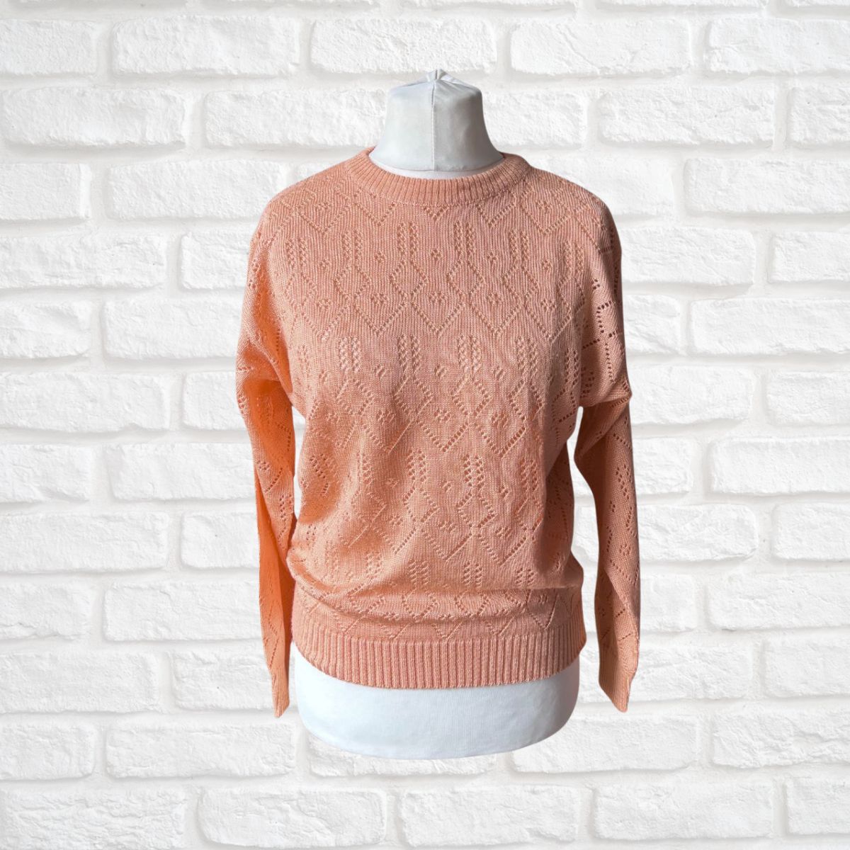 A peach-colored sweater displayed on a white mannequin torso against a white brick wall background.