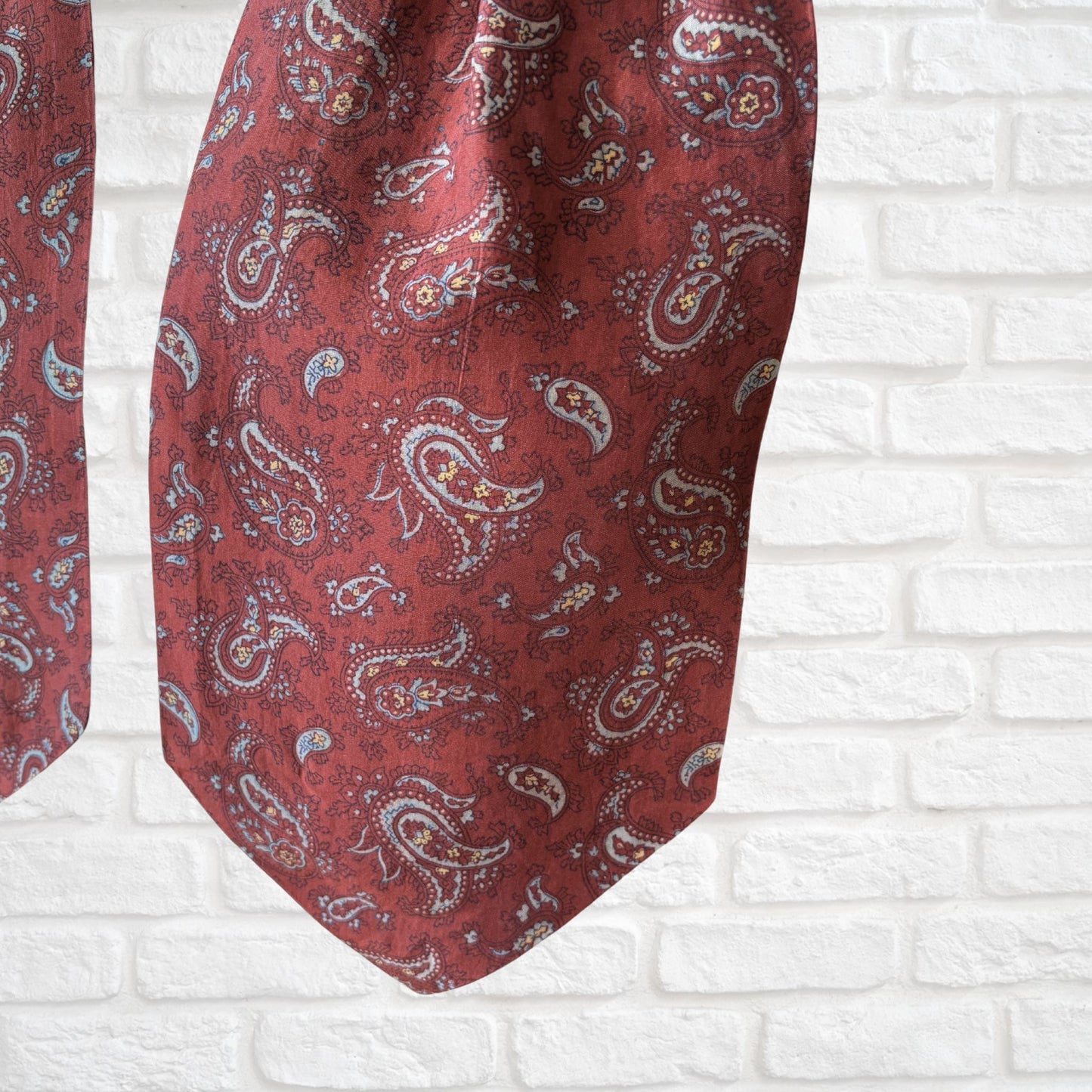 A vintage silk cravat featuring a paisley print in red, grey, yellow, and blue, displayed on a mannequin against a white brick wall background.