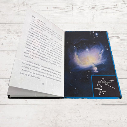 An open book lying on a white wood surface, showing a page with text on the left and a space-themed cover or interior spread on the right with a dark starry nebula illustration and a small constellation diagram.
