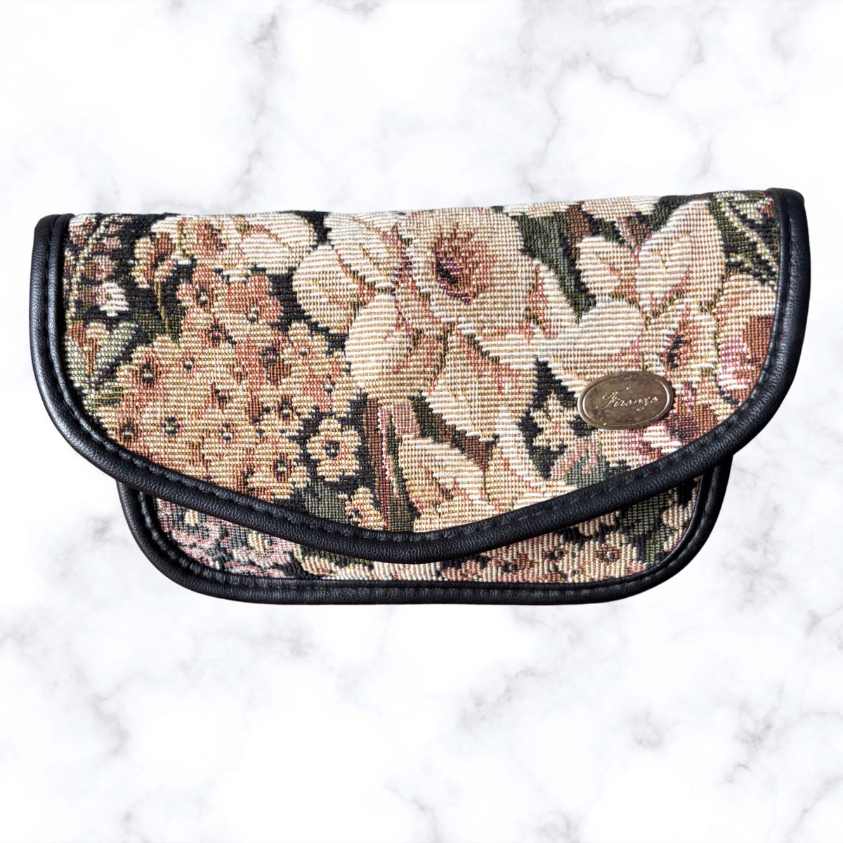 A vintage floral tapestry pouch with black leather trim, featuring a small oval metal brand plaque 