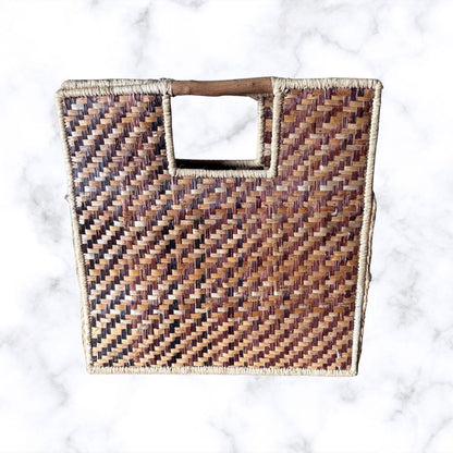 A square woven straw tote bag with a single cutout handle, resting on a white marble surface.