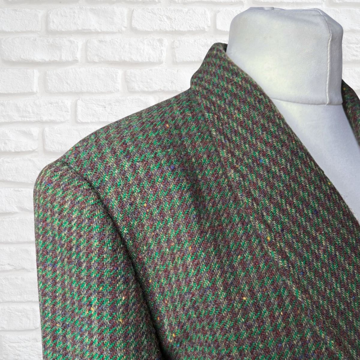 A close-up of a tweed blazer draped on a white mannequin torso, featuring a green and brown houndstooth pattern with subtle gold flecks, set against a white brick wall background.