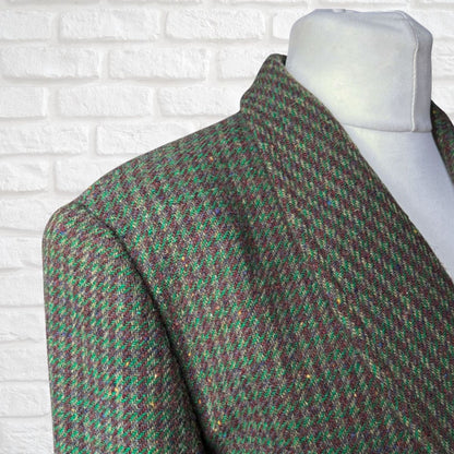 A close-up of a tweed blazer draped on a white mannequin torso, featuring a green and brown houndstooth pattern with subtle gold flecks, set against a white brick wall background.