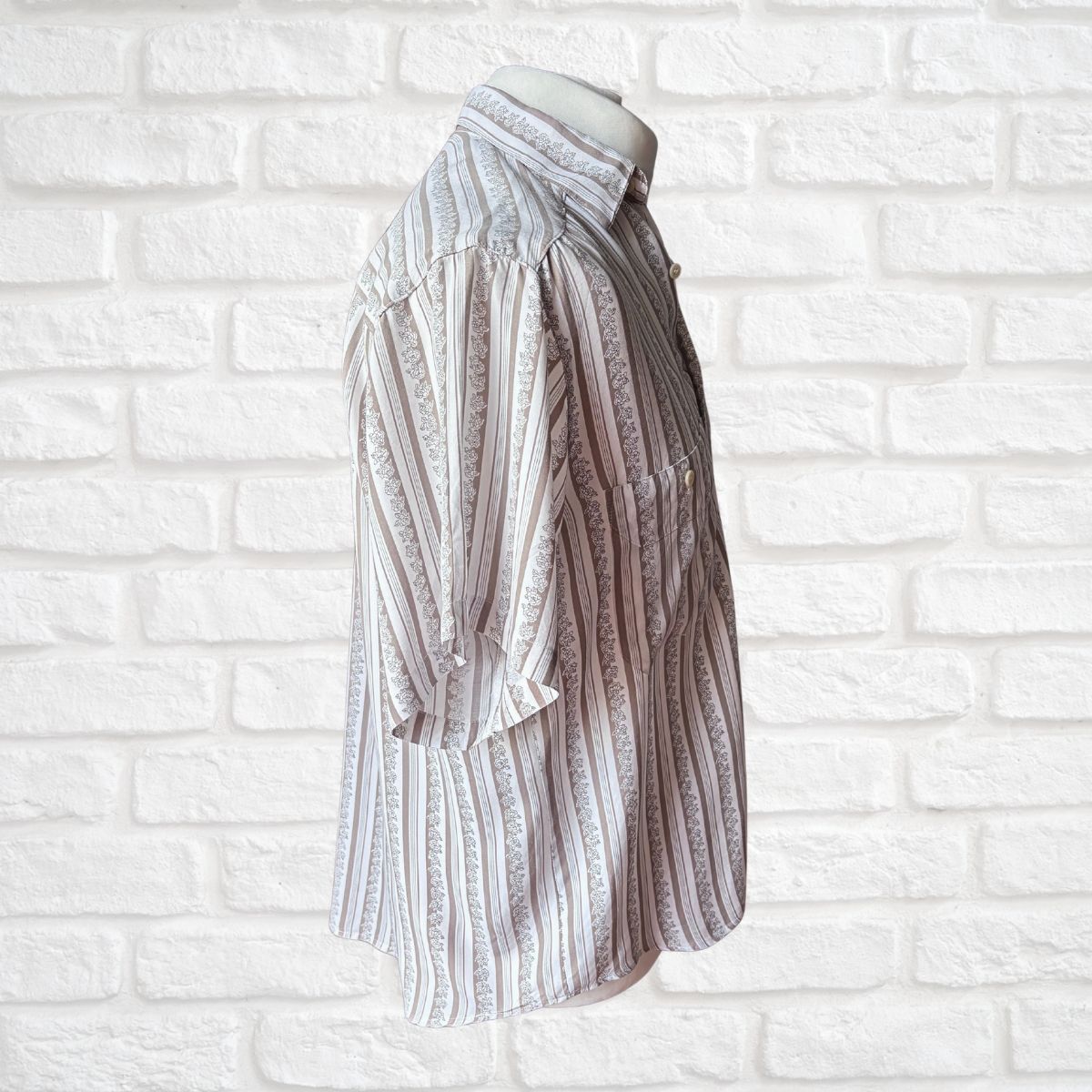 A long-sleeve button-up shirt with vertical stripes in beige, brown, and white, featuring a subtle floral pattern, displayed on a mannequin torso against a white brick wall.