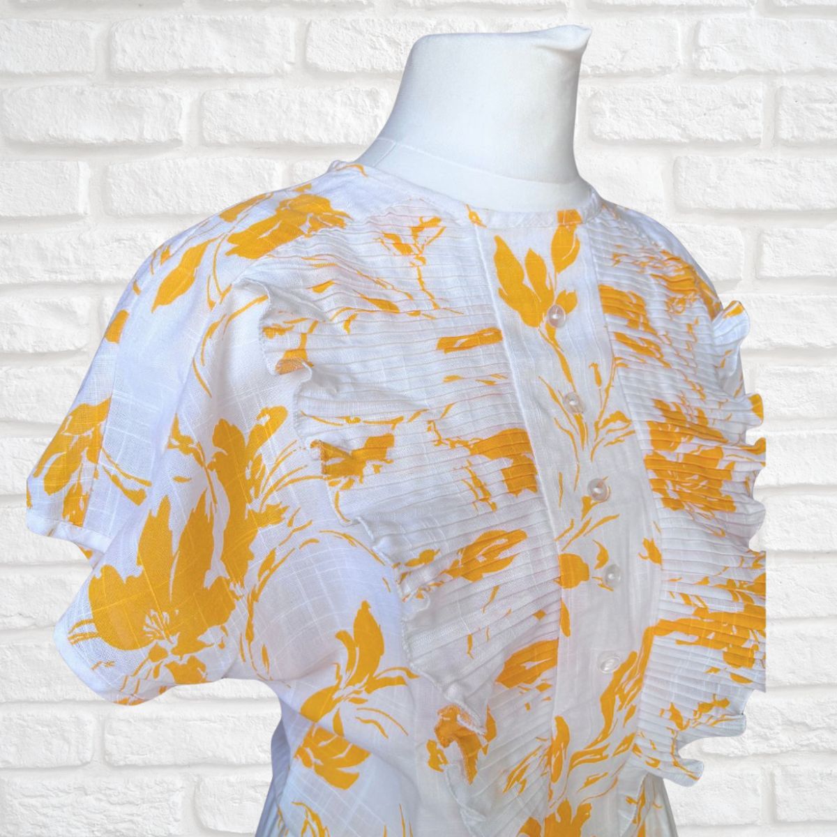 White dress with yellow floral patterns on a mannequin against a white brick wall.