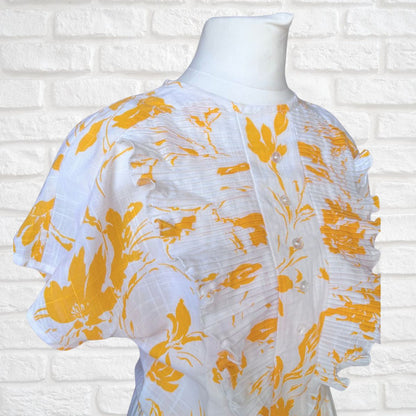 White dress with yellow floral patterns on a mannequin against a white brick wall.