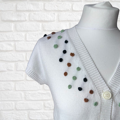 White knit cardigan sweater with short sleeves and colorful pom-pom embellishments, displayed on a white mannequin torso against a white brick wall background.