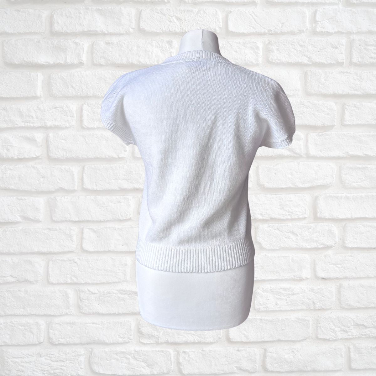 A white short-sleeve knit top displayed on a white dress form mannequin, shown from the back against a white painted brick wall background.