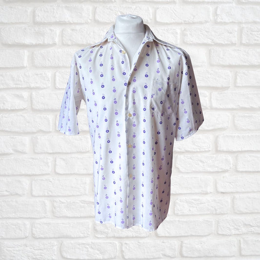 A white short-sleeve button-up shirt with a purple and blue floral pattern, displayed on a white mannequin torso against a white brick wall background.
